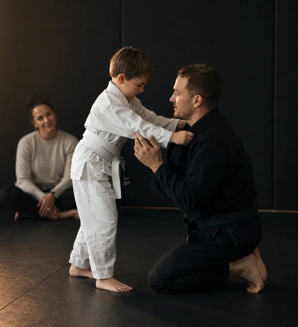 A coach kneeling to adjust a young white belt's gi while a parent watches