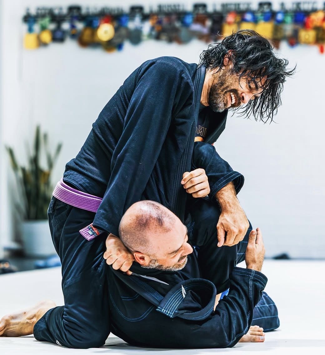 Matt McAlexander in a navy gi and purple belt, controlling a training partner from top position during a roll